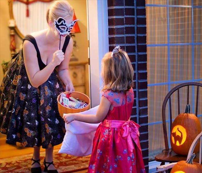 Setting is a front porch. A woman wearing a mask holding a basket of candy for a little girl in a red costume.